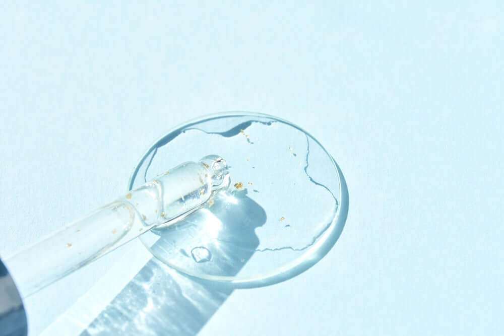 Close-up of a glass dropper with a clear liquid and tiny gold flecks, resting on a light blue surface with water reflections.