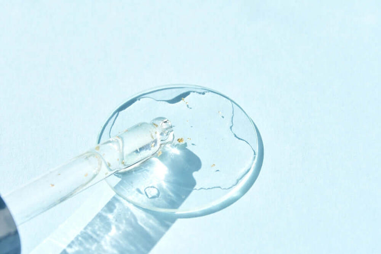 Close-up of a glass dropper with a clear liquid and tiny gold flecks, resting on a light blue surface with water reflections.