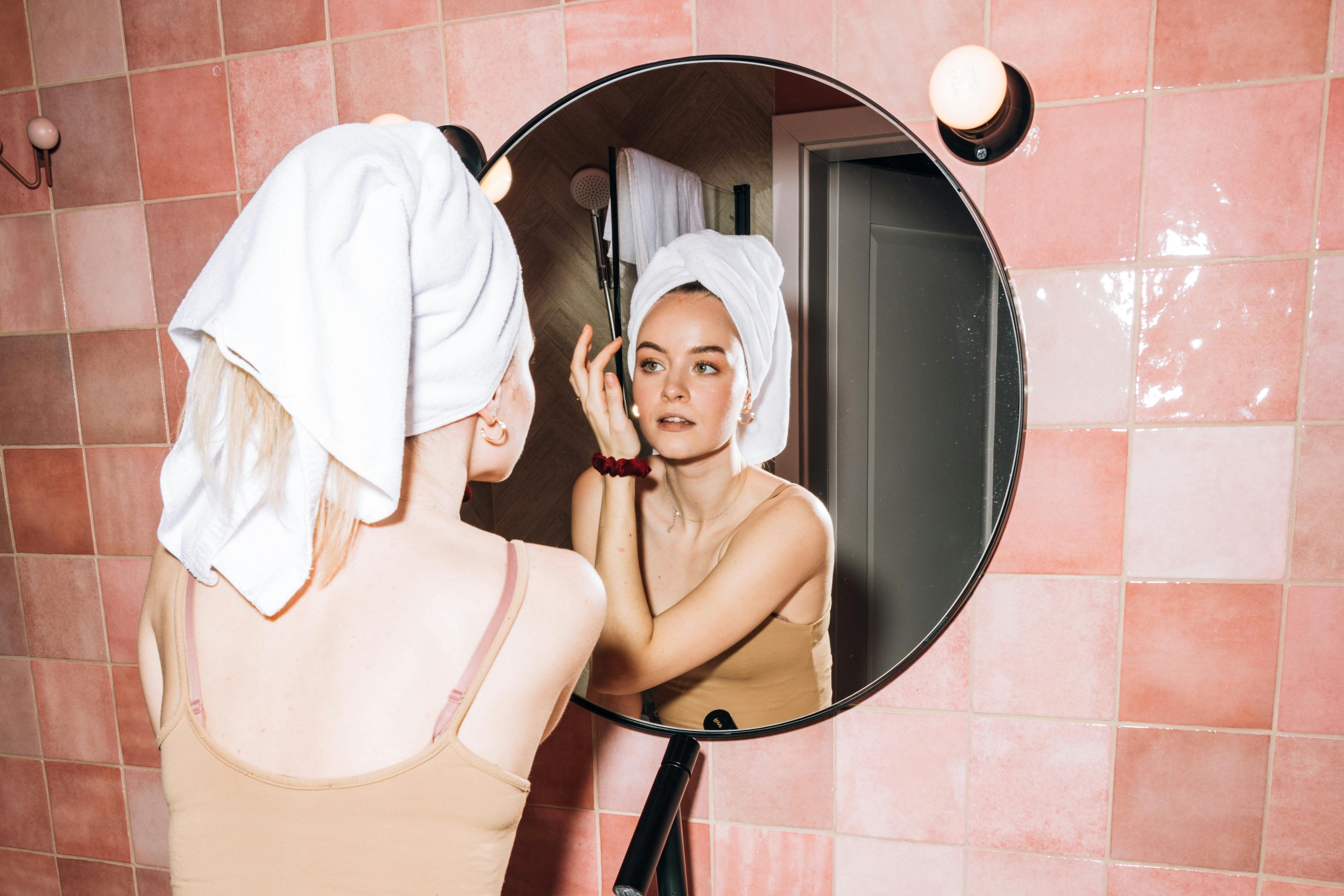 Young woman with a bath towel wrapped around her head, standing in front of a bathroom mirror. She is looking closely at her skin with a focused expression, inspecting her face. The bathroom has soft lighting, with pink tones on the walls.